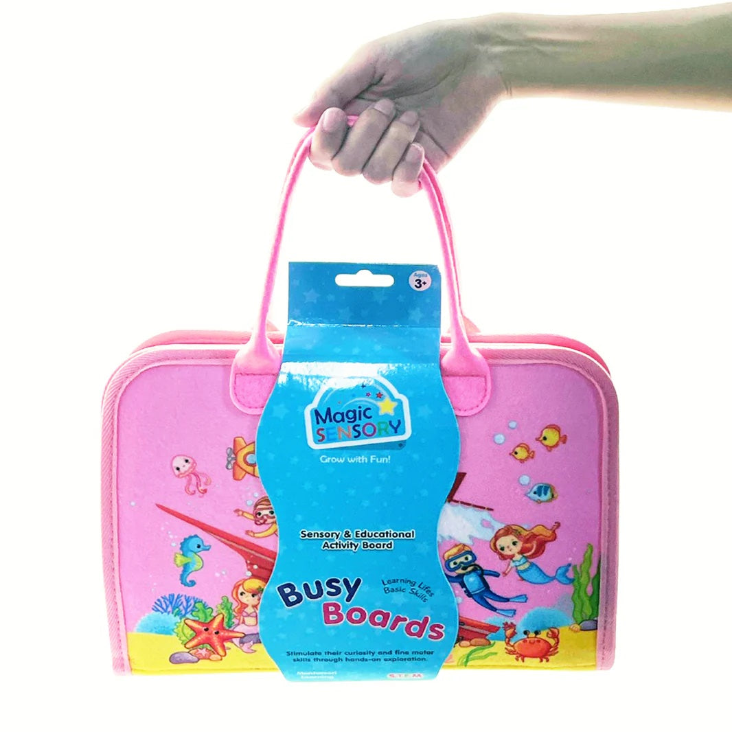 Pink activity board with 'Magic Seas & Magic Busy Boards' packaging held by a hand on a white background