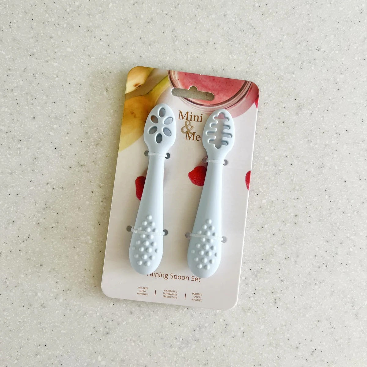 Two baby spoons on a packaging card with 'Mini & Me' branding on a textured surface.