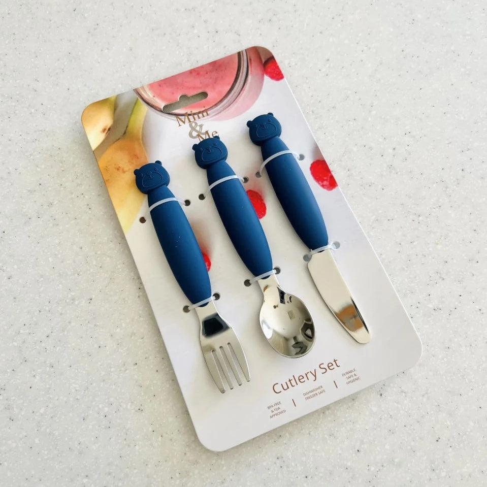 A three-piece baby cutlery set in its original retail packaging, featuring blue silicone handles and silver stainless steel utensils (fork, spoon, knife) on a speckled light gray countertop.