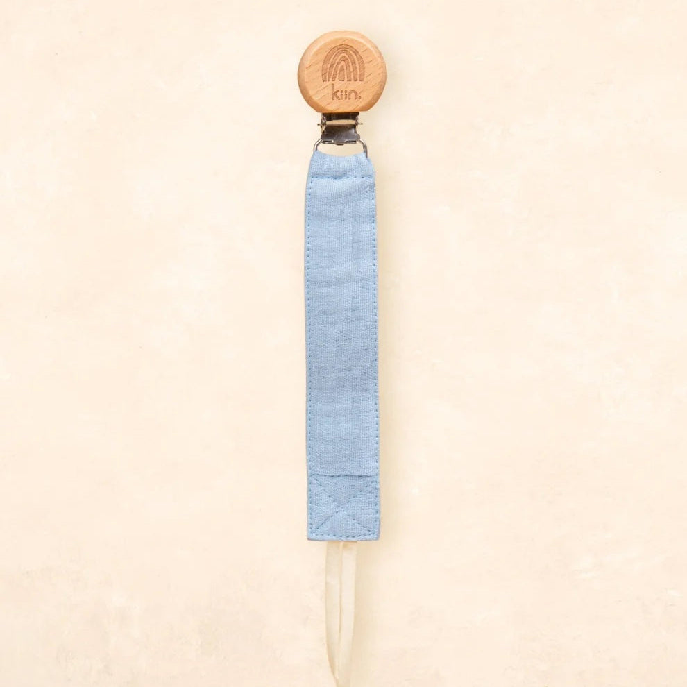 A light blue fabric strap accessory with a round natural wood end and a small metal connector, shown against a warm beige textured background.