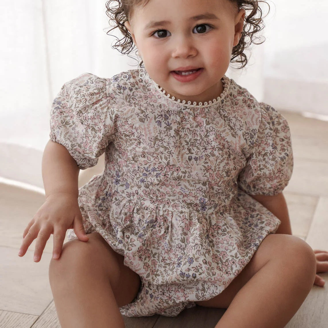 Child wearing a floral romper sitting on a light wooden floor.