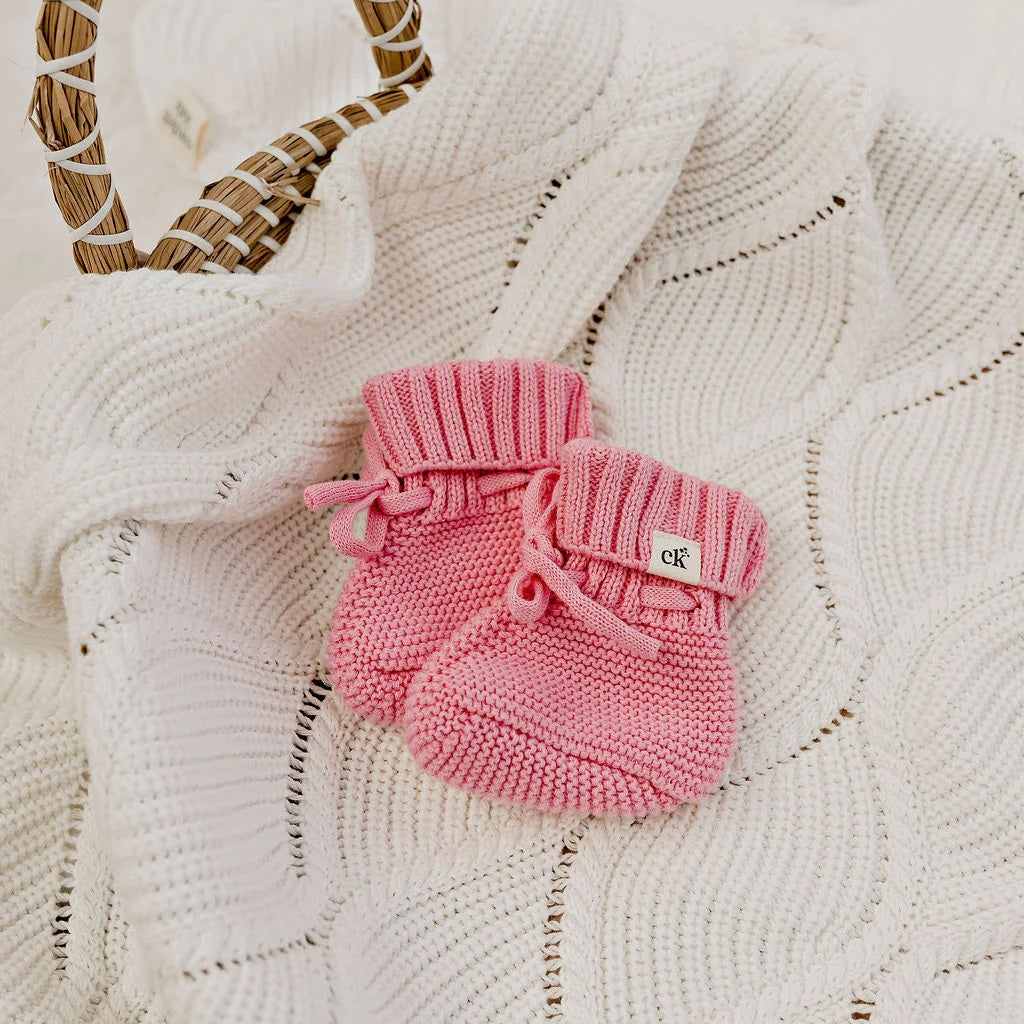 Pink knitted baby booties on a textured white surface with a woven heart decoration.