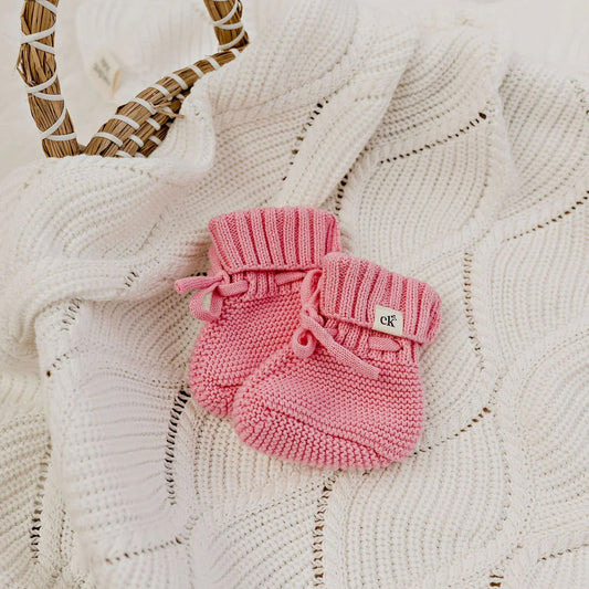 Pink knitted baby booties on a textured white surface with a woven heart decoration.