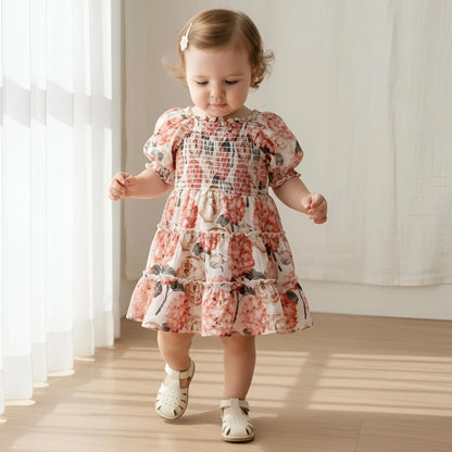 A toddler girl wearing a short puff-sleeve tiered dress with a pink and beige floral print, walking indoors on a wooden floor near sheer white curtains.