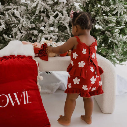 Child in a red dress with white floral patterns standing near a decorated Christmas tree.