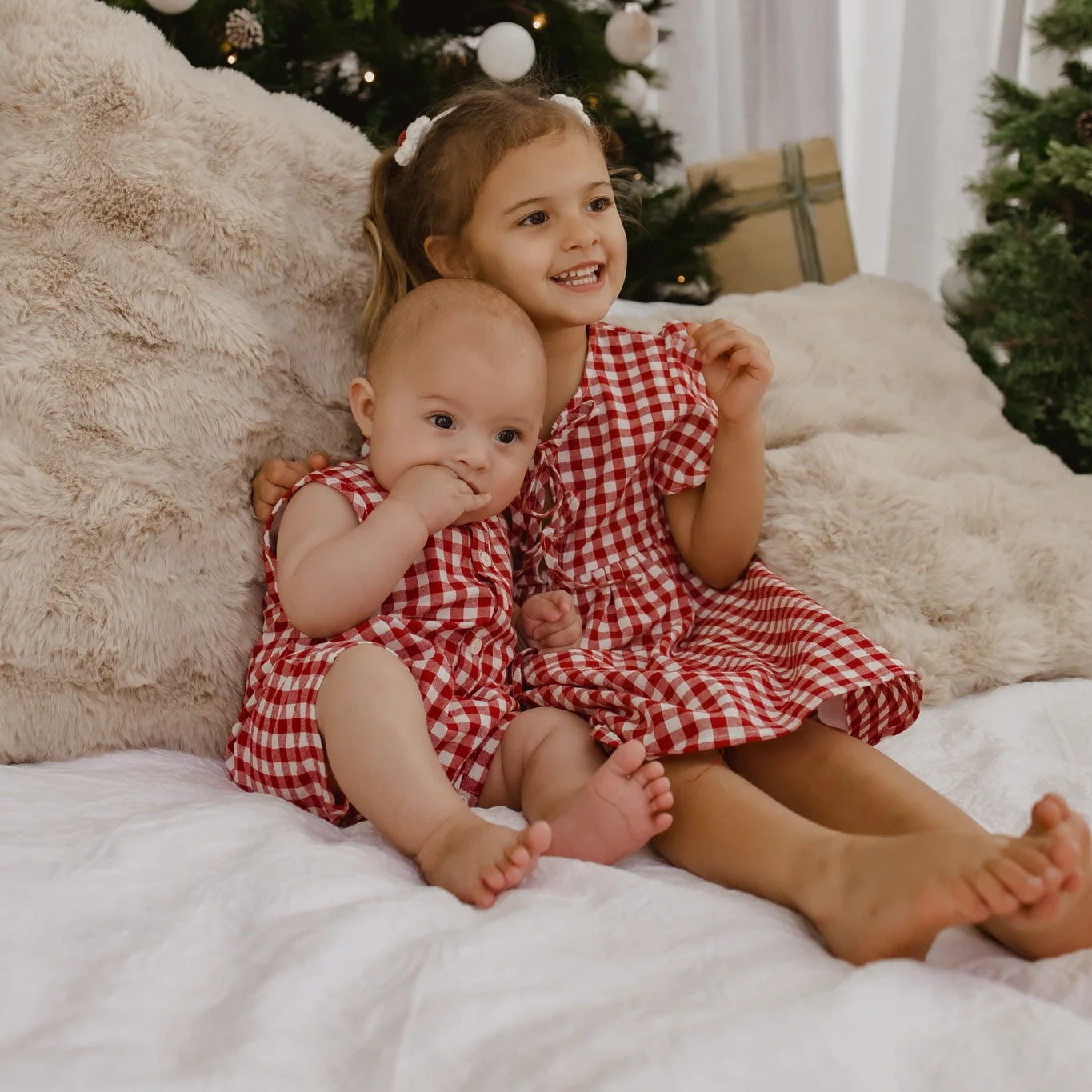 Two young girls in red checkered dresses sitting on a bed with Christmas trees in the background.