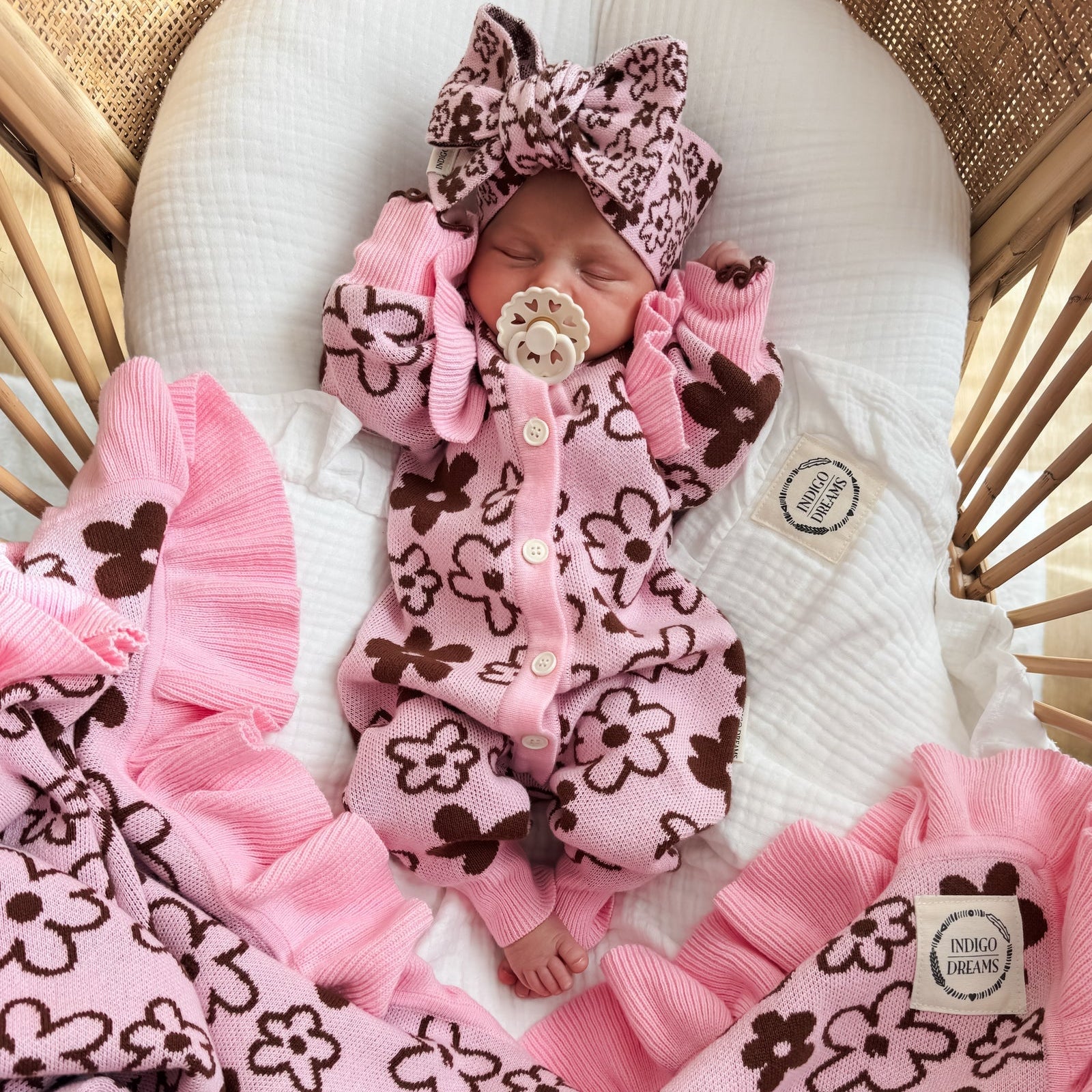 Newborn baby in pink and brown outfit with matching headband and blanket in a crib.