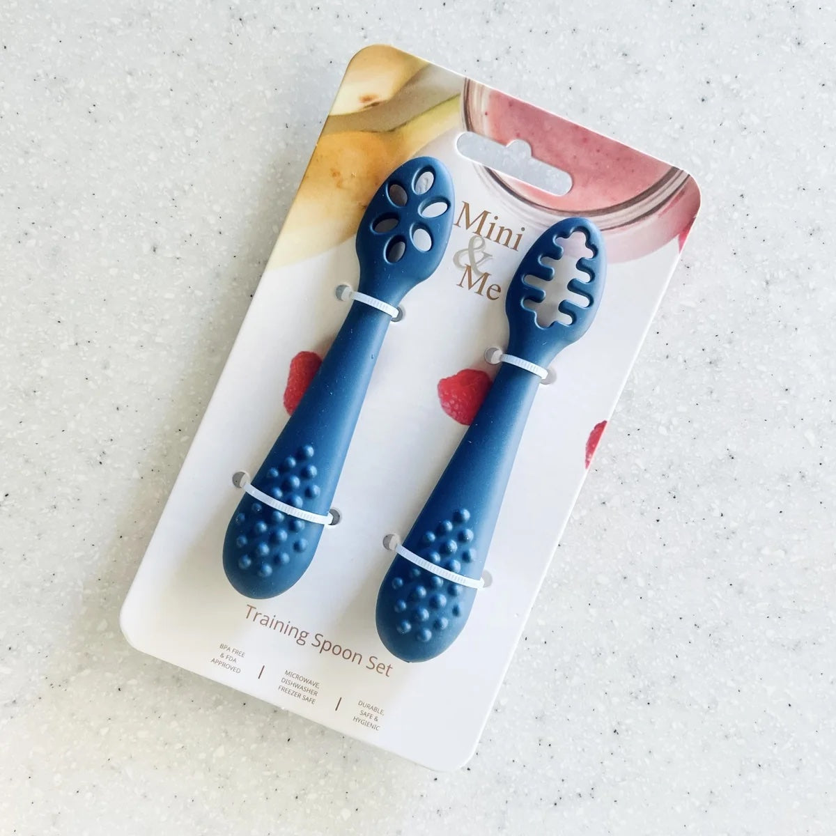 A blue training spoon set in its original retail packaging, displayed on a light speckled countertop. The set includes two small silicone training spoons with textured handles and cutout heads, labeled 'Mini & Me Training Spoon Set'.