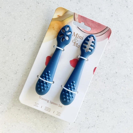 A blue training spoon set in its original retail packaging, displayed on a light speckled countertop. The set includes two small silicone training spoons with textured handles and cutout heads, labeled 'Mini & Me Training Spoon Set'.
