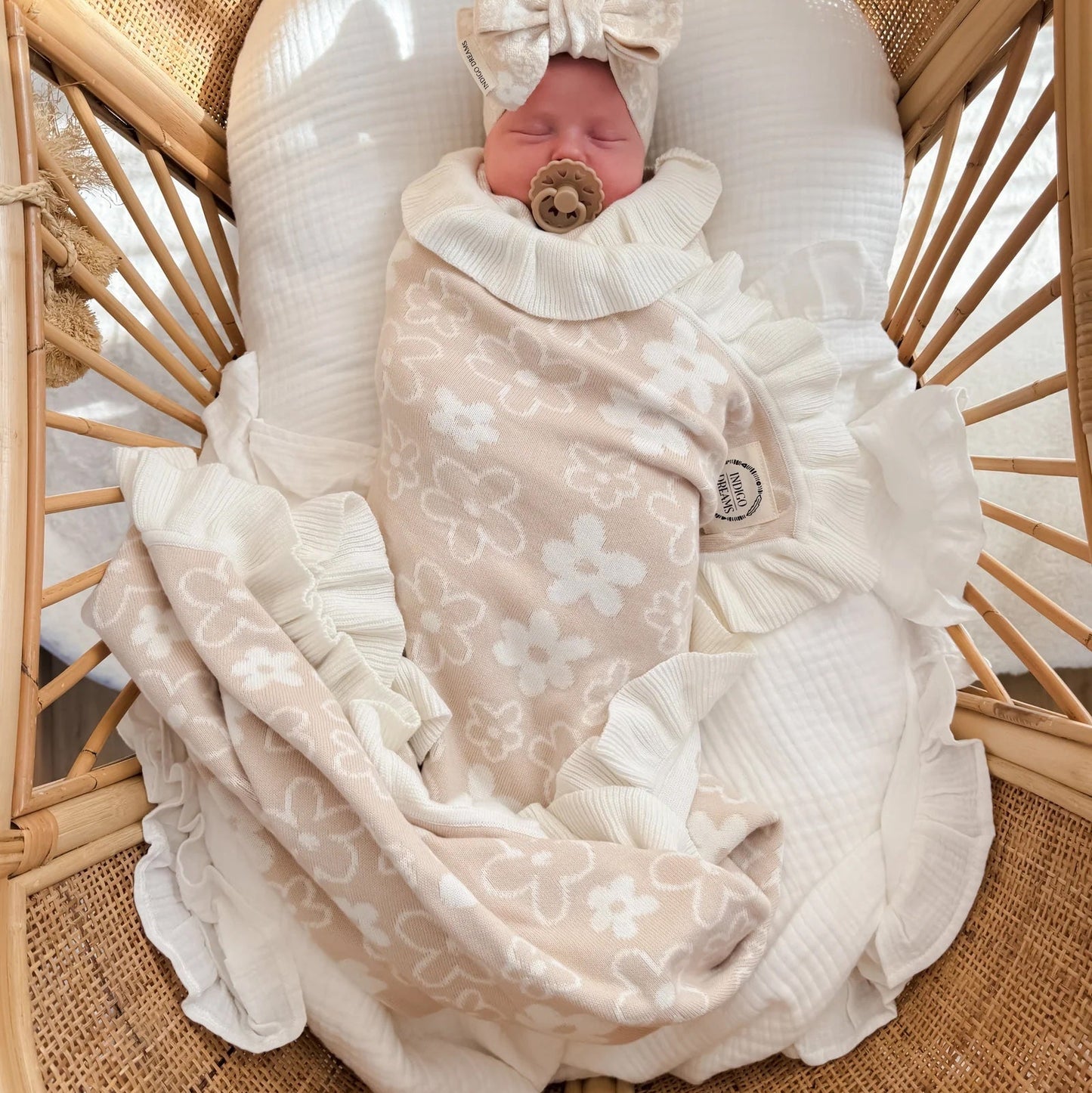 A newborn baby is sleeping peacefully in a woven rattan bassinet, swaddled in a soft beige blanket with white floral patterns and a matching ruffled bonnet.