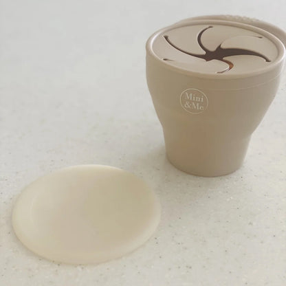 A beige reusable cup with a matching lid placed beside it on a light speckled surface. The cup has a printed logo reading 'Mini & Me' and a dark abstract graphic on the lid.