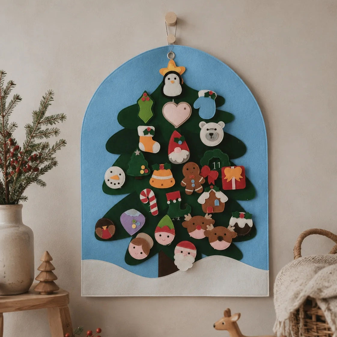 A wooden Christmas advent tree wall hanging with colorful felt ornaments, displayed on a beige wall in a styled room setting with a wooden toy deer, basket, and dried flowers.