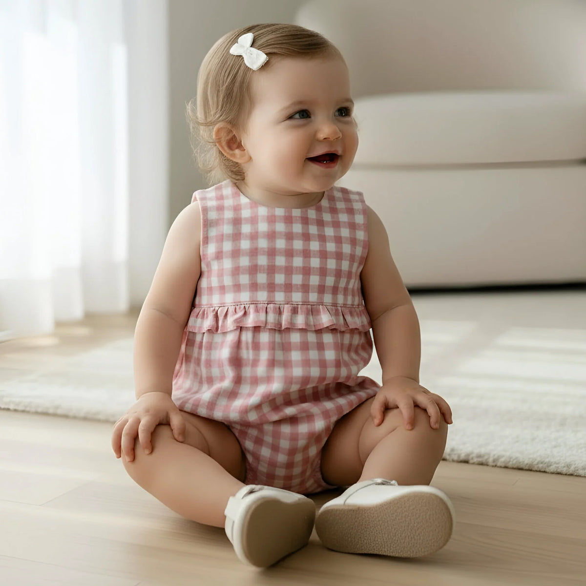 A baby girl sitting on a light wood floor in a bright room, wearing a pink and white gingham sleeveless romper with a ruffle at the waist and light beige shoes.