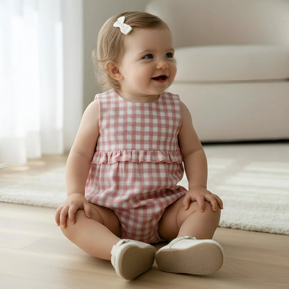 A baby girl sitting on a light wood floor in a bright room, wearing a pink and white gingham sleeveless romper with a ruffle at the waist and light beige shoes.