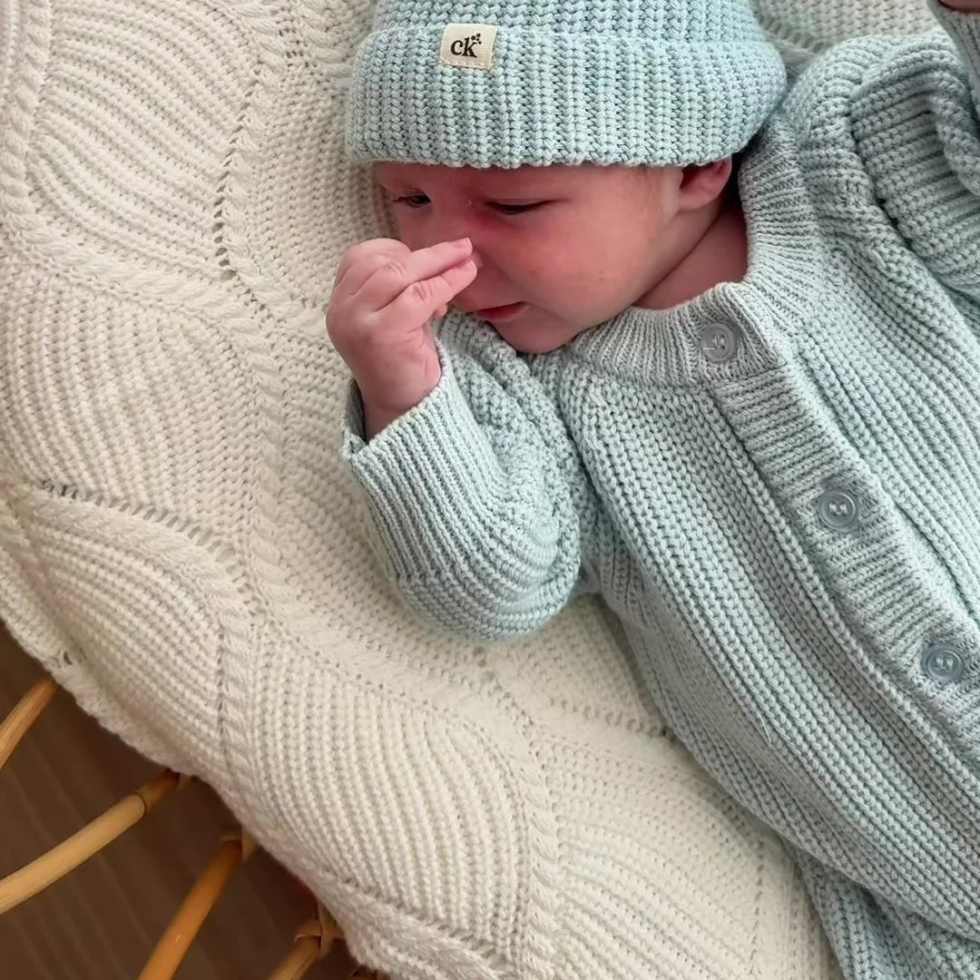 Newborn baby in a light blue knitted outfit and hat lying on a textured white cushion.