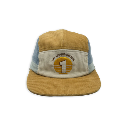 A five-panel trucker hat made of tan corduroy with light blue mesh side panels and a cream front panel. The front features an embroidered circular patch with a large number 1 and the text 'I'M AROUND THE WORLD'.