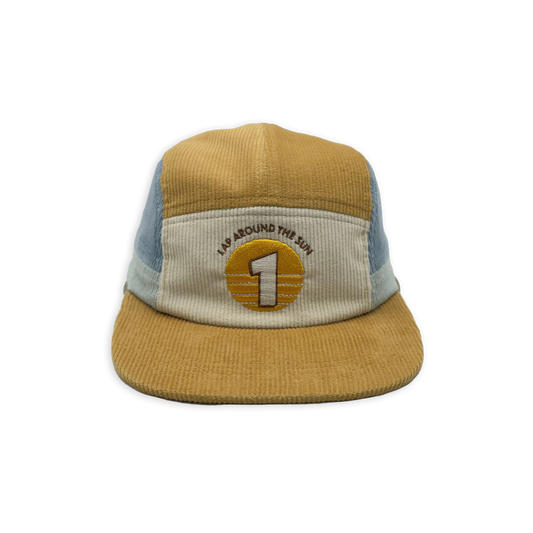 A five-panel trucker hat made of tan corduroy with light blue mesh side panels and a cream front panel. The front features an embroidered circular patch with a large number 1 and the text 'I'M AROUND THE WORLD'.