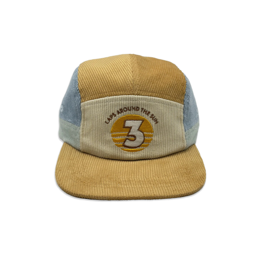 A 5-panel trucker cap made from mixed materials, featuring a mustard yellow corduroy front panel and brim, light blue mesh side panels, and a beige front patch with an embroidered circular logo.