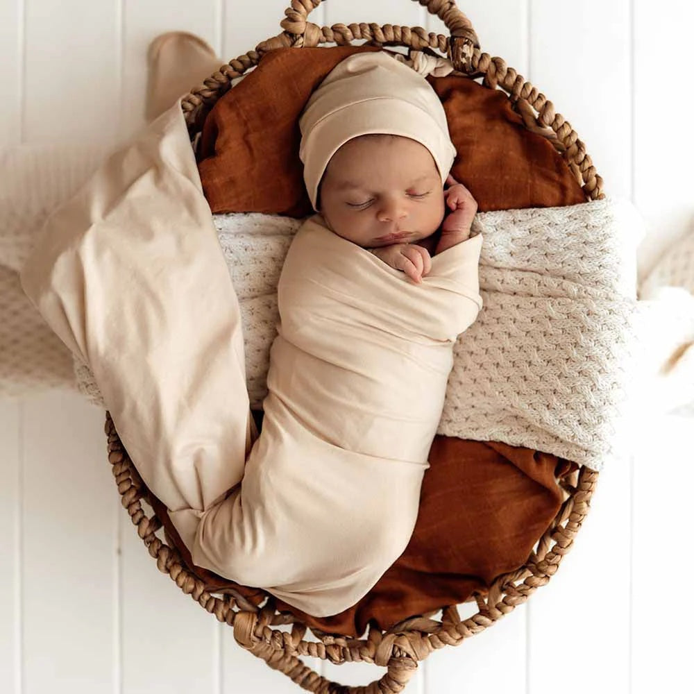Newborn baby wrapped in beige swaddle and cap, lying in a woven basket with brown and white blankets.