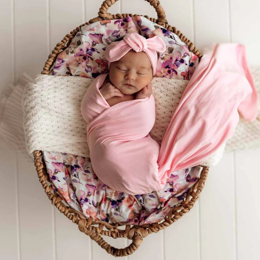 Newborn baby wrapped in pink fabric with a floral headband, lying in a woven basket.