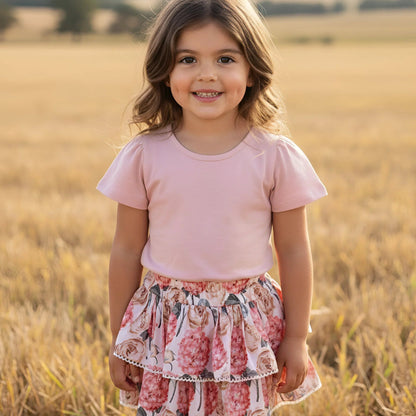 A young girl with light brown hair stands in a golden wheat field, wearing a light pink short-sleeve t-shirt and a tiered ruffled skirt with a pink, red, and white floral pattern.