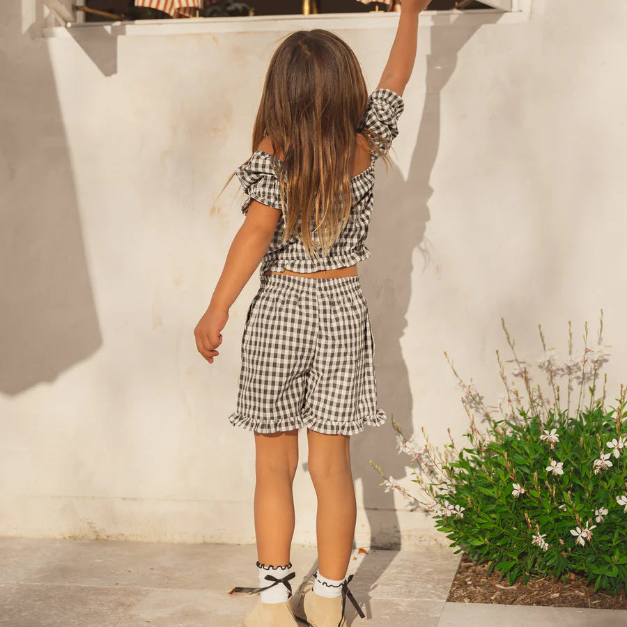 Young girl in a checkered outfit standing in front of an open window with curtains.