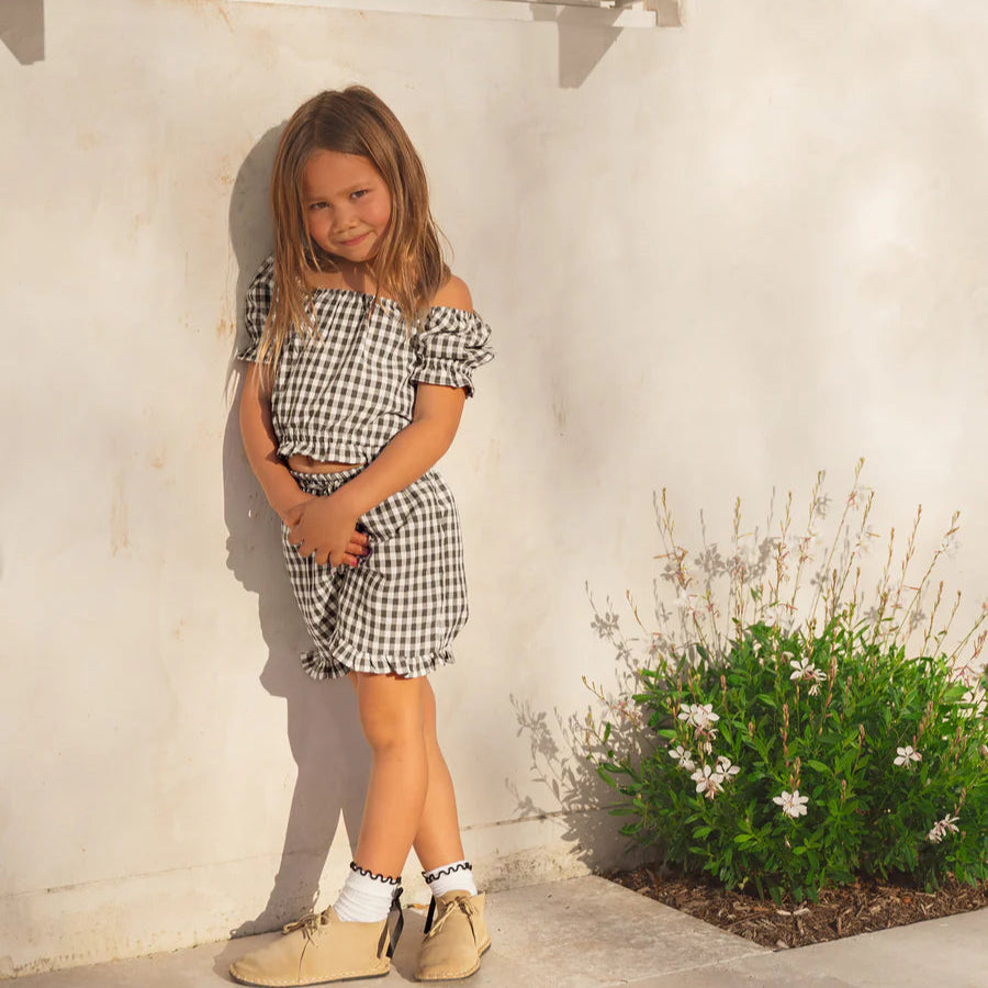 Young girl in a checkered dress standing against a white wall with a window and plant.