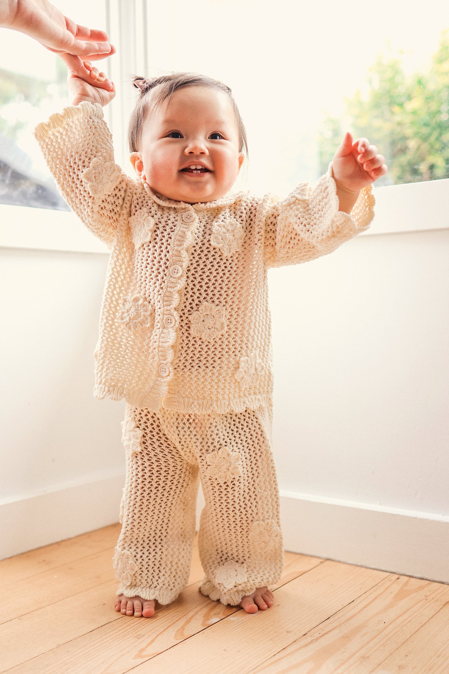 Baby in a beige knitted outfit standing on a wooden floor with a blurred background