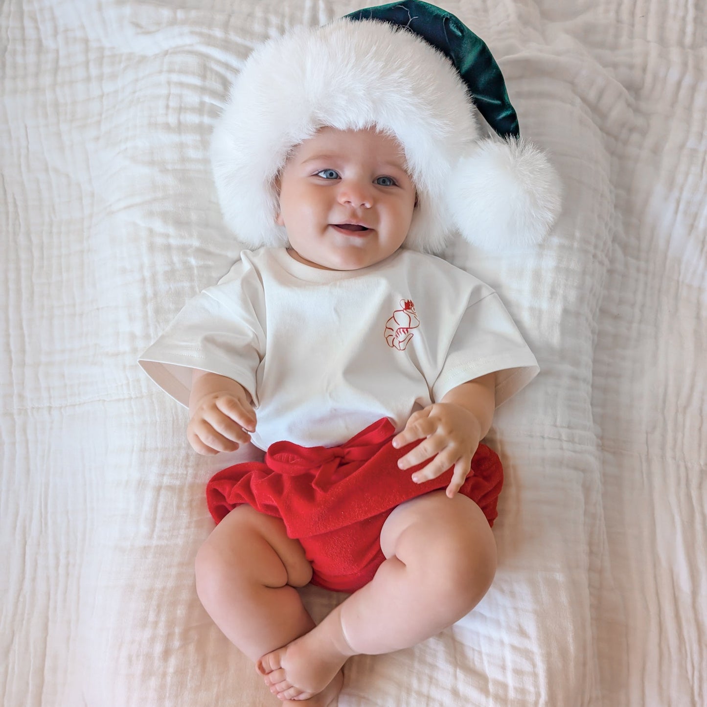 Baby wearing a white shirt with a red diaper, sitting on a white textured surface.