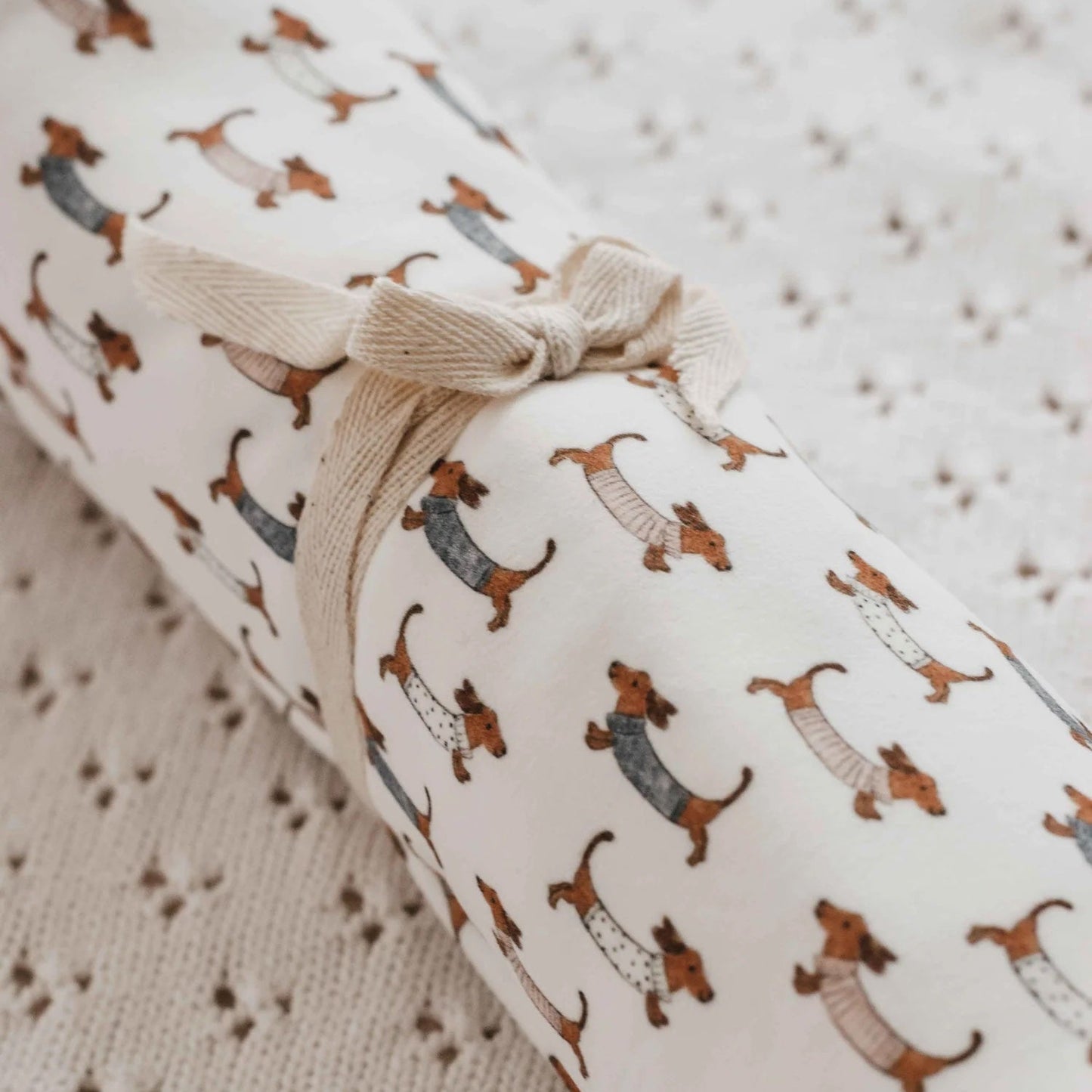 A rolled fabric item, likely a blanket or throw, featuring a dachshund dog pattern in brown and gray on a cream background, tied with a beige ribbon. It is laid on a textured cream knit blanket.