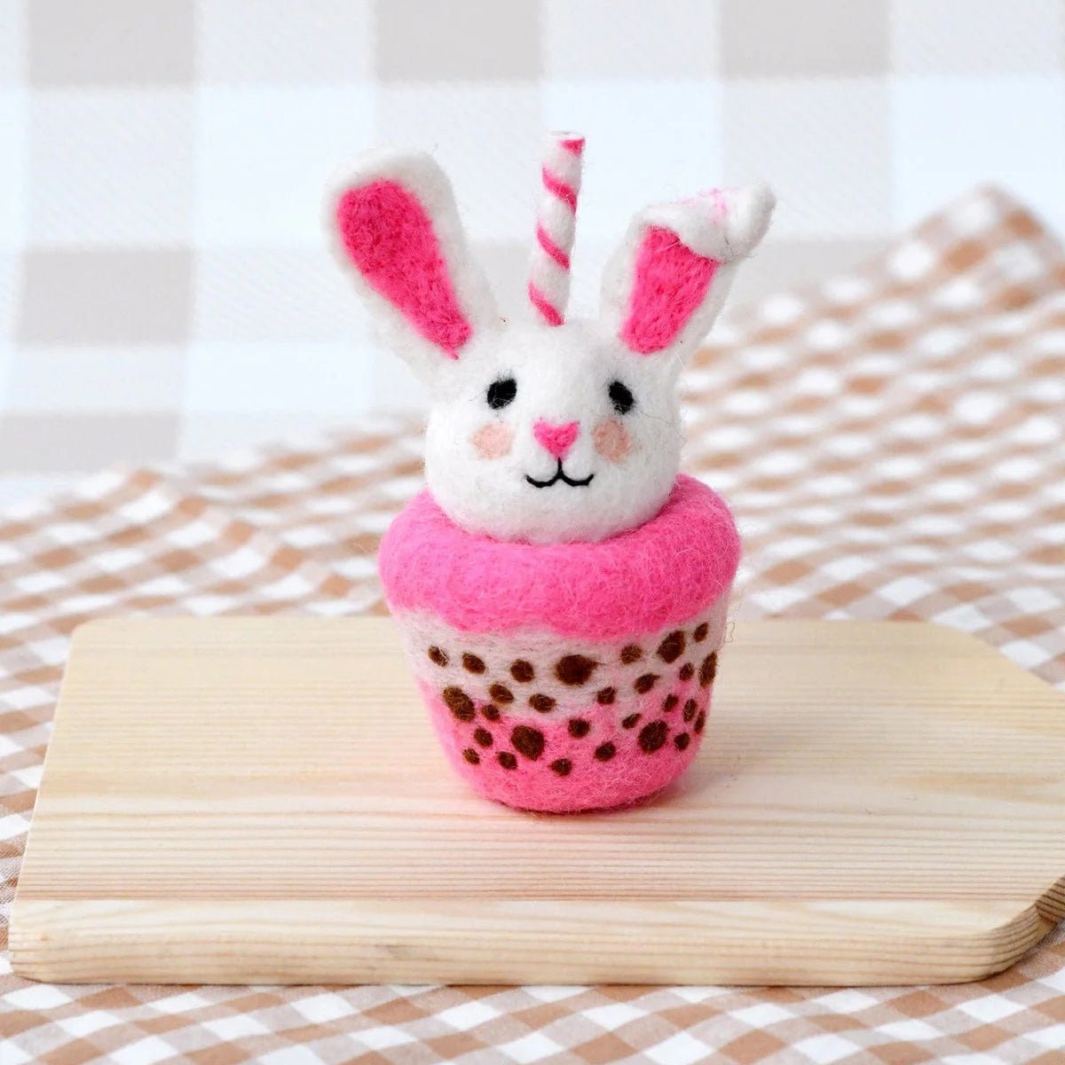 Plush toy rabbit in a pink cupcake on a wooden surface with a checkered fabric background