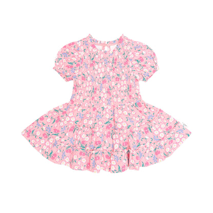 A pink short-sleeve dress with a dense floral and abstract print in pink, purple, and green, featuring a smocked bodice and ruffled hem, shown flat on a white background.