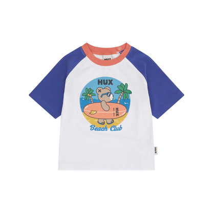A children's short-sleeve raglan t-shirt by HUX, featuring a white body, royal blue sleeves, and a coral orange collar. The front is decorated with a colorful cartoon graphic of a bear on a beach with the text 'HUX Beach Club'.