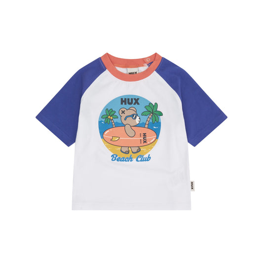 A children's short-sleeve raglan t-shirt by HUX, featuring a white body, royal blue sleeves, and a coral orange collar. The front is decorated with a colorful cartoon graphic of a bear on a beach with the text 'HUX Beach Club'.