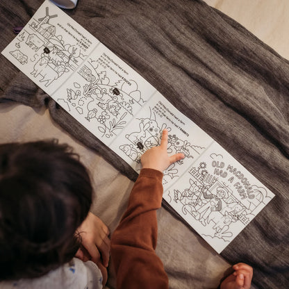 Child coloring a picture of a farm scene with black outlines on a white sheet, sitting on a brown blanket.