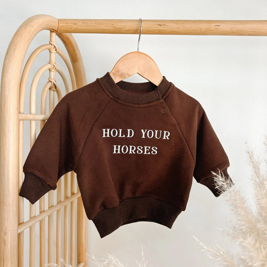 Brown sweatshirt with 'Hold Your Horses' text on a hanger against a neutral background