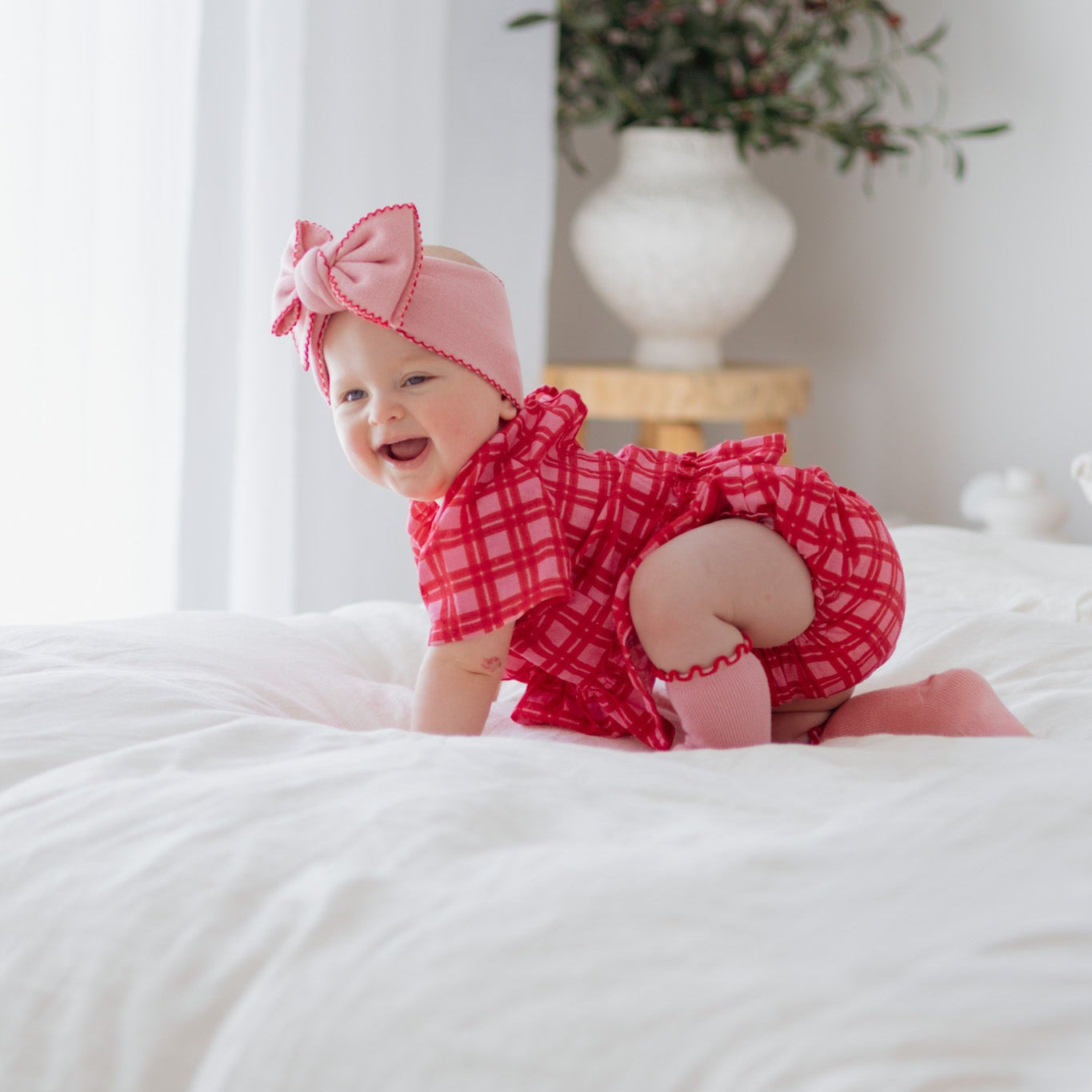 Baby in a red outfit and headband sitting on a white bed with a plant in the background.