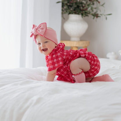 Baby in a red outfit and headband sitting on a white bed with a plant in the background.