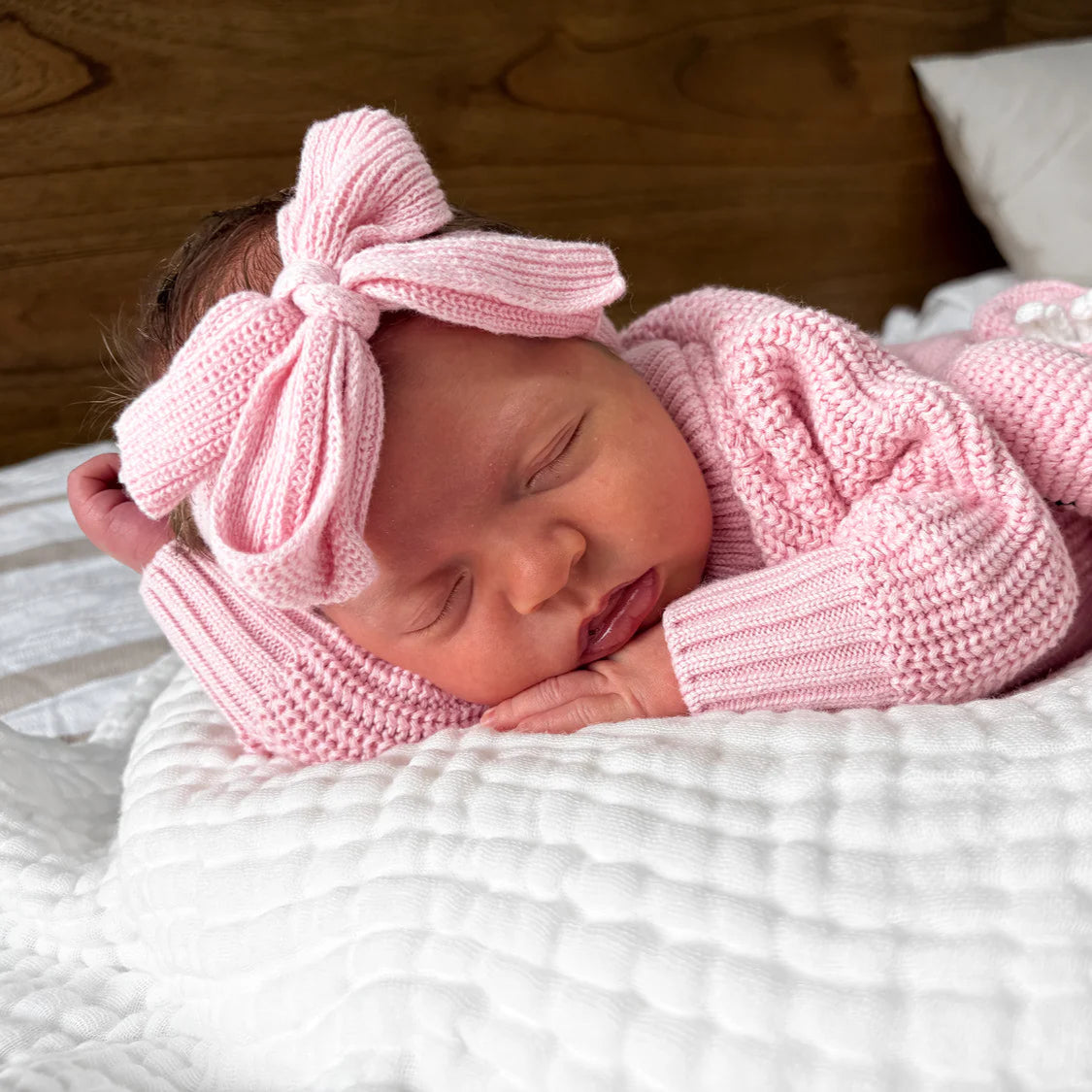 Newborn baby in pink knitted outfit with bow headband on white blanket