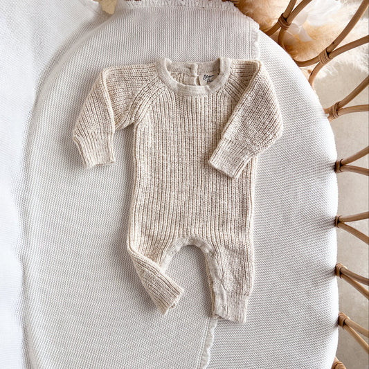 Knitted baby romper on a textured surface with a wicker chair in the background