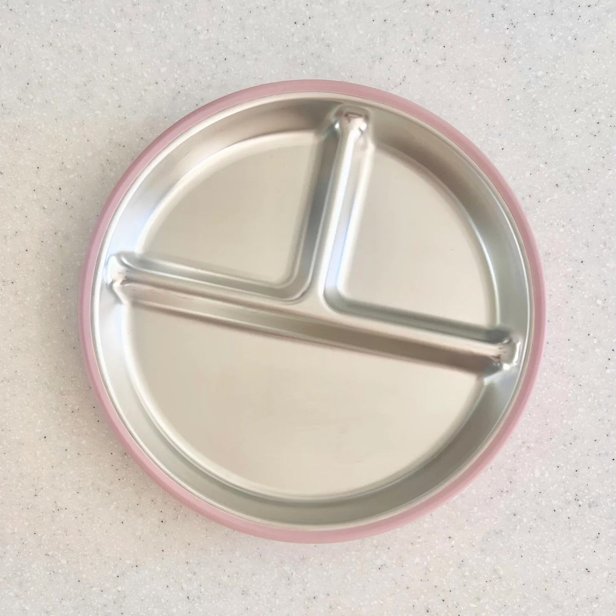 A round stainless steel divided baby plate with a pink rim, shown from a top-down view on a speckled light gray countertop.