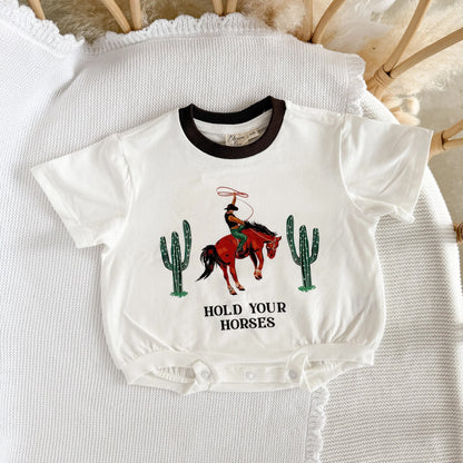 White baby onesie with cowboy graphic and text 'Hold Your Horses' on a textured surface.