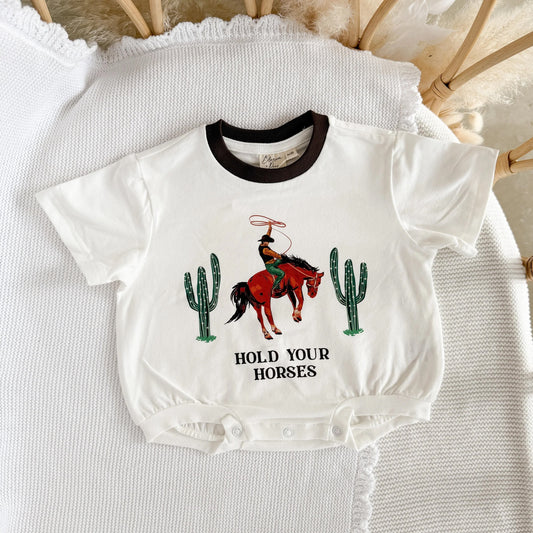 White baby onesie with cowboy graphic and text 'Hold Your Horses' on a textured surface.