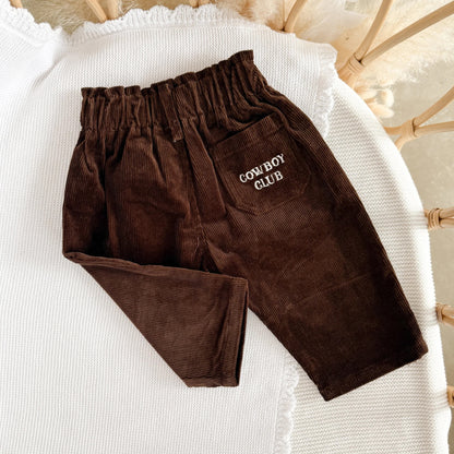 Brown corduroy pants with 'Cowboy Club' embroidery on a white surface.