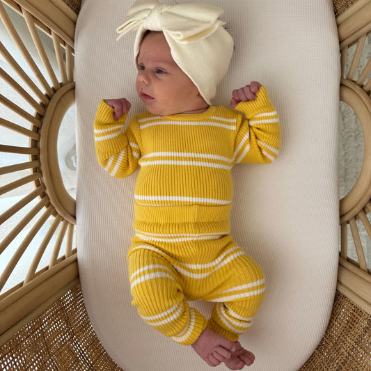Newborn baby in a yellow outfit and white hat lying on a white oval object with a wicker background.