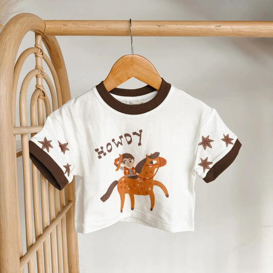 Children's t-shirt with cowboy design and 'HOWDY' text on a wooden hanger.