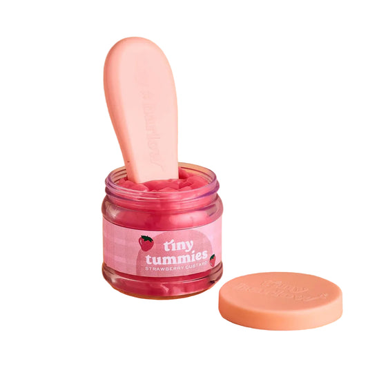 A pink strawberry gummy jar by tiny tummies, featuring a clear jar filled with pink gummies, a pink scoop tool, and a peach-colored lid on a solid beige background.