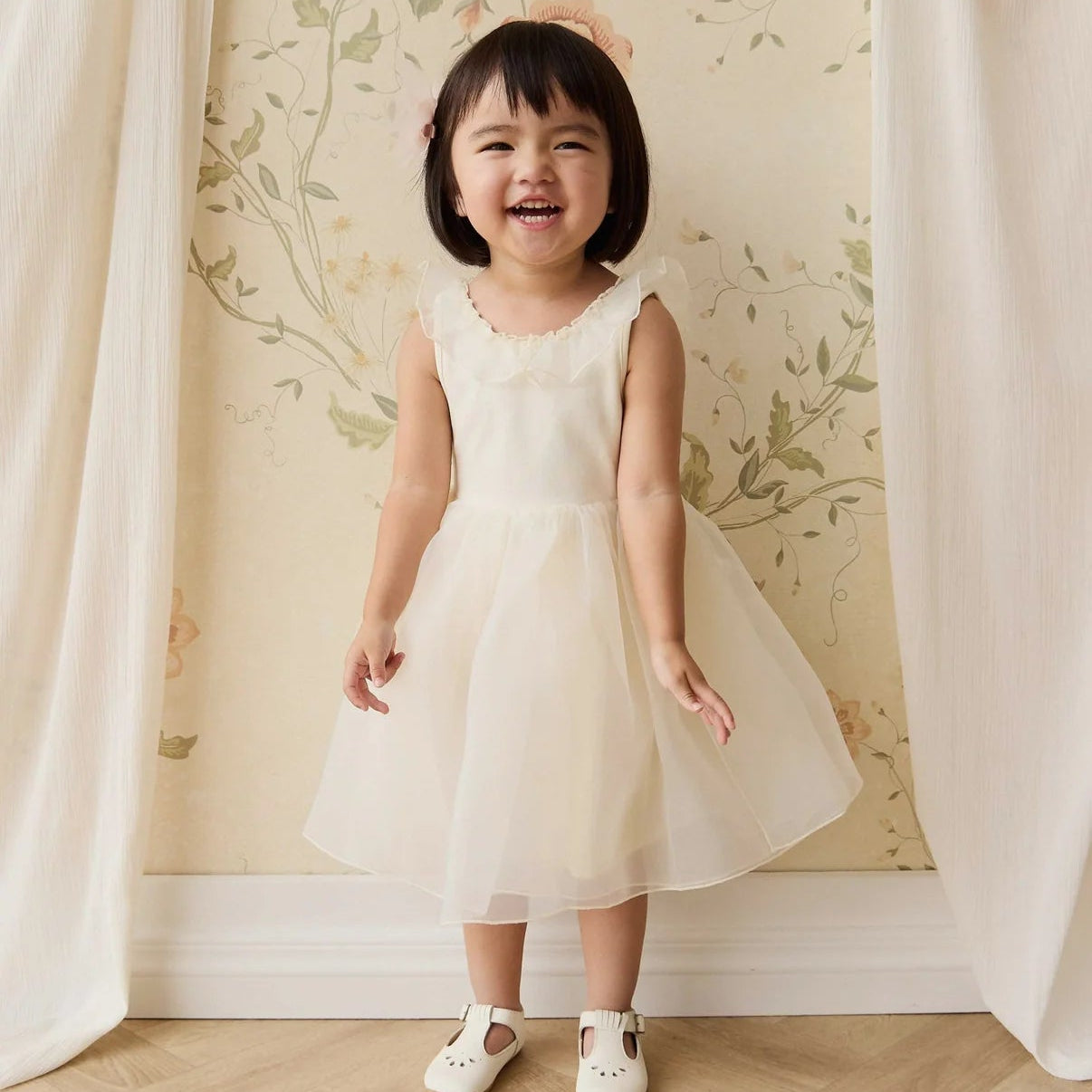 Young girl in a white dress standing in front of a floral wallpapered wall.