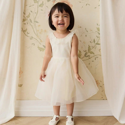 Young girl in a white dress standing in front of a floral wallpapered wall.