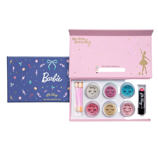 A Barbie-themed makeup set shown with its blue illustrated outer box and the open pink inner kit. The kit contains six small round cosmetic pots with smiling face designs, two pink-handled applicators, and a black lipstick tube labeled 'Be little Shine-by'.